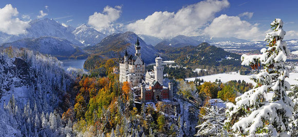 A beautiful landscape of Neuschwanstein Castle nestled among the Alps, covered in snow with colorful autumn foliage surrounding it. The majestic castle towers over the lush hills, framed by mountains and a clear blue sky adorned with white clouds, creating a serene and picturesque scene perfect for a jigsaw puzzle.