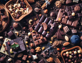 A beautifully arranged flat lay of assorted chocolates and nuts. The image features various types of chocolate bars, chocolate-covered nuts, and decorative elements like mint leaves and wooden bowls filled with mixed nuts. The rich brown tones contrast against the wooden background, creating a mouthwatering visual appeal reminiscent of a dessert paradise.
