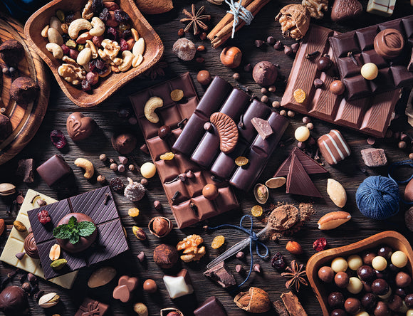 A beautifully arranged flat lay of assorted chocolates and nuts. The image features various types of chocolate bars, chocolate-covered nuts, and decorative elements like mint leaves and wooden bowls filled with mixed nuts. The rich brown tones contrast against the wooden background, creating a mouthwatering visual appeal reminiscent of a dessert paradise.