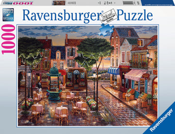 A Ravensburger jigsaw puzzle titled 'Cafe Scene' featuring a charming street lined with cafes, shops, and tables under twinkling lights. The setting captures a vibrant evening atmosphere with intricate details, inviting viewers to piece together a picturesque outdoor scene reminiscent of a cozy European cafe.