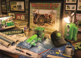 A detailed work desk featuring John Deere memorabilia, including a vibrant green jigsaw puzzle of a tractor, blueprints, a mug, and various decorative items. The background showcases framed artwork, a computer displaying a John Deere image, and a desk lamp illuminating the workspace.