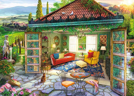 A vibrant jigsaw puzzle titled 'Tuscan Oasis' by Ravensburger, featuring a cozy room with a red sofa and yellow chair, surrounded by lush green landscapes, colorful flowers, and a stone pathway. The scene captures the warmth and charm of a Tuscan retreat, with decorative elements like a chandelier and plants enhancing its inviting ambiance.