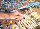 A hand holding a blue jigsaw puzzle piece above a partially completed jigsaw puzzle depicting a scenic view of Rome. Surrounding the puzzle are multiple scattered pieces, showcasing vibrant colors and intricate designs. The table surface is wooden, providing a warm backdrop for the game.