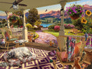 A picturesque front porch scene depicted in a large jigsaw puzzle by Ravensburger. The vibrant image features a cozy seating area with a dog resting on a pet bed, colorful potted plants, and a beautiful view of mountains and a lake in the background. A cat curiously observes from a nearby chair, while a decorative rug adds charm to the space. The overall atmosphere is tranquil and inviting, highlighting a serene outdoor living environment.