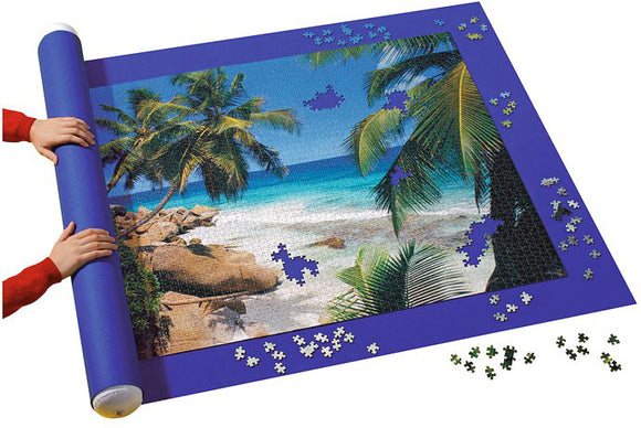 The Giant Puzzle Stow & Go! by Ravensburger is a jigsaw puzzle in progress, featuring a vibrant beach scene with palm trees and blue ocean. A person is rolling up the puzzle on a mat designed to keep pieces secure, while scattered puzzle pieces are visible around the edges.