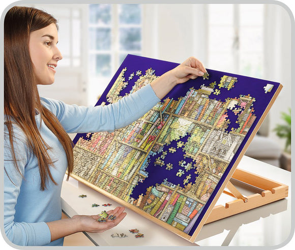 A young woman is piecing together a colorful jigsaw puzzle on a wooden puzzle board. The puzzle features an intricate design of bookshelves and various images, displayed on a blue background. She smiles as she connects pieces, with several jigsaw pieces scattered on the table below.