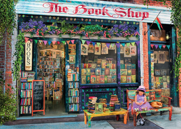 A vibrant jigsaw puzzle depicting a charming bookshop exterior. The shop features a sign that reads 'The Book Shop' with colorful flowers and bunting. Books fill the shelves and a chalkboard advertises a book sale. A young girl in a pink hat sits on a colorful bench with a cat beside her and a small dog on a leash, creating a whimsical, inviting atmosphere.