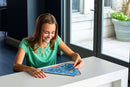 A young girl sitting at a white table, playing with the Triazzles Dolphins mechanical puzzle by Think Fun. She has a joyful expression as she interacts with the colorful puzzle featuring dolphins, surrounded by a bright and modern living space.