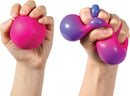 A hand holding the NeeDoh Color Change toy, which is a stress-relief fidget ball. The toy is bright pink, soft and squeezable, stretching and changing colors as it is manipulated. It appears in two different shapes, illustrating its pliability and engaging visual features.