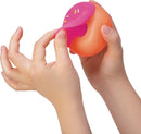 A pair of hands holding a NeeDoh Dohnuts stress ball. The toy is pink and orange and resembles a donut, featuring colorful sprinkles on the top. It's being squeezed, showcasing its stretchy, squishy texture, perfect for stress relief and sensory play.