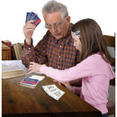 A grandfather and his granddaughter are sitting at a wooden table playing Quiddler, a word card game by Set Games. The grandfather holds three Quiddler cards in his hand while the girl, focused and engaged, moves cards on the table, illustrating a moment of fun, family interaction, and learning through play.