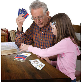 A grandfather and his granddaughter are sitting at a wooden table playing Quiddler, a word card game by Set Games. The grandfather holds three Quiddler cards in his hand while the girl, focused and engaged, moves cards on the table, illustrating a moment of fun, family interaction, and learning through play.