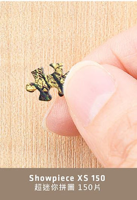 A close-up image of a hand holding a single jigsaw puzzle piece. The piece is intricately designed with a colorful pattern that includes shades of green and gold. This image represents the Puppies in a Gift Box XS 150 pc Puzzle by Pintoo, showcasing the quality and detail of its pieces.