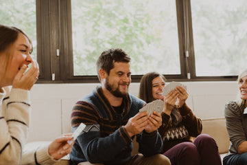 A group of four friends joyfully playing Momo's Shanghai Rummy, a card game made by Solid Roots. They are sitting together, laughing and engaging with their cards in a cozy indoor setting, surrounded by sunlight filtering through the windows.