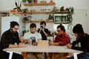 A group of five people seated around a wooden table, each focused on their smartphones. The scene is set in a bright kitchen filled with plants and kitchenware. A wooden Device Detox Box is at the center of the table, labeled 'Phones? Let's put them where we're going.' The atmosphere suggests a social gathering with a hint of digital detox.