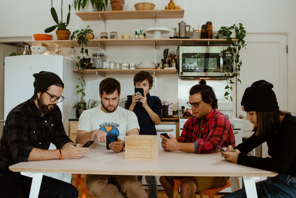 A group of five people seated around a wooden table, each focused on their smartphones. The scene is set in a bright kitchen filled with plants and kitchenware. A wooden Device Detox Box is at the center of the table, labeled 'Phones? Let's put them where we're going.' The atmosphere suggests a social gathering with a hint of digital detox.