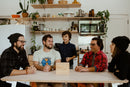 A group of five adults and a child gathered around a table in a cozy kitchen. They are engaged in conversation, with various kitchen items and plants in the background. In the forefront, a wooden box labeled 'Phones? Where we’re going, we don’t need phones' is placed on the table, suggesting a focus on disconnecting from technology and enjoying quality time together.