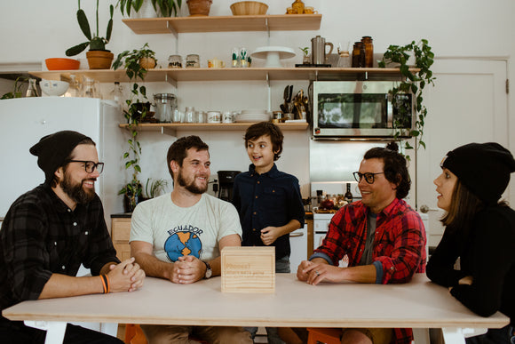 A group of five adults and a child gathered around a table in a cozy kitchen. They are engaged in conversation, with various kitchen items and plants in the background. In the forefront, a wooden box labeled 'Phones? Where we’re going, we don’t need phones' is placed on the table, suggesting a focus on disconnecting from technology and enjoying quality time together.