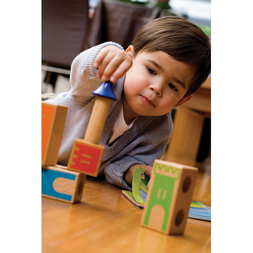 A young boy is playing with Castle Logix, a mechanical puzzle by SmartGames. He is intently stacking colorful wooden blocks, featuring designs of castles and towers. The boy, dressed in a grey sweater, is focused on balancing a blue turret atop a block, while other blocks are spread out on the wooden floor, showcasing vibrant, engaging graphics that stimulate creativity and cognitive skills.