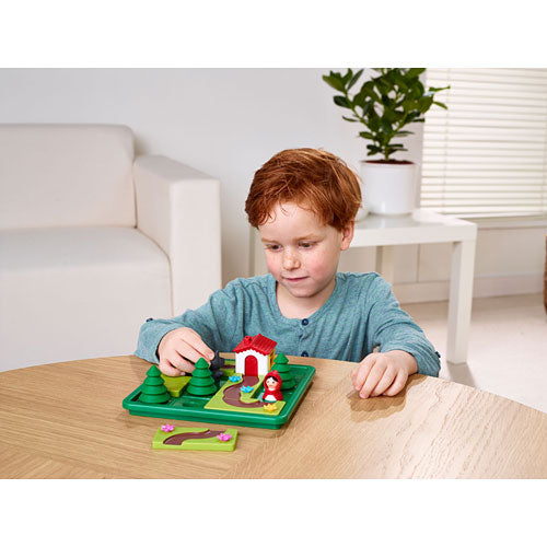 A young child playing with the Little Red Riding Hood Deluxe mechanical puzzle from SmartGames. The set features a colorful forest scene with green trees, a red-roofed house, and the character of Little Red Riding Hood. The child is focusing on assembling the components on a wooden table in a bright room.