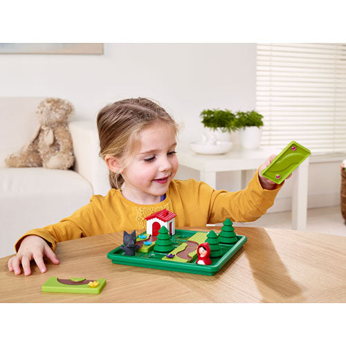 A young girl plays with the Little Red Riding Hood Deluxe mechanical puzzle by SmartGames. The colorful game features whimsical components including trees, a house, a wolf, and a figurine of Little Red Riding Hood on a green base. The child looks engaged as she manipulates pieces of the puzzle, showcasing creativity and problem-solving skills.