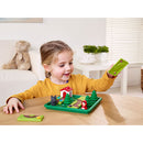 A young girl plays with the Little Red Riding Hood Deluxe mechanical puzzle by SmartGames. The colorful game features whimsical components including trees, a house, a wolf, and a figurine of Little Red Riding Hood on a green base. The child looks engaged as she manipulates pieces of the puzzle, showcasing creativity and problem-solving skills.