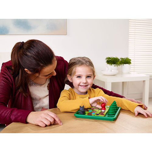 A joyful young girl and an adult engage in play with the Little Red Riding Hood Deluxe mechanical puzzle by SmartGames. The colorful board features charming characters and trees, encouraging creative thinking and problem-solving skills. The girl smiles as she manipulates the pieces, showcasing a fun and interactive learning experience.