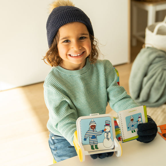 A smiling child wearing a navy beanie and mint green sweater holds a colorful two-sided challenge toy that features a snowman and two figures in winter clothes. The toy encourages engagement and problem-solving skills, with a spiral notebook for solutions. The setting is bright and cheerful, perfect for creative play.