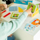 A child interacting with the Dress Code mechanical puzzle by Smart Toys & Games. The child is pointing at a vibrant illustrated card featuring a character holding a flower. Various colorful transparent cards and challenge books are spread across the table, showcasing the engaging and educational nature of the puzzle.