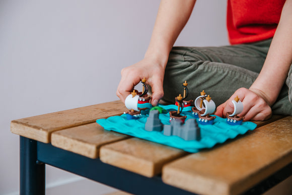 A child plays with the Pirates Crossfire mechanical puzzle by Smart Toys and Games. The game features colorful pirate ships navigating through blue waves, adding a fun and engaging element to playtime.