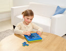 A young girl is engaged in playing the Jungle Hide & Seek Mechanical Puzzle by SmartGames. She focuses intently on manipulating colorful puzzle pieces on a blue and green puzzle board set on a wooden table. A cozy living room setting with a white couch and a blue cushion is visible in the background.