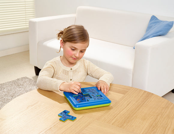 A young girl is engaged in playing the Jungle Hide & Seek Mechanical Puzzle by SmartGames. She focuses intently on manipulating colorful puzzle pieces on a blue and green puzzle board set on a wooden table. A cozy living room setting with a white couch and a blue cushion is visible in the background.