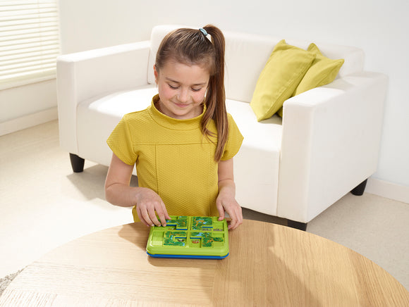 A young girl wearing a yellow shirt is playing with the Jungle Hide & Seek mechanical puzzle by SmartGames. The puzzle features a colorful design with animals on tiles, providing an interactive and engaging experience on a wooden table, with a bright white sofa and green pillows in the background.