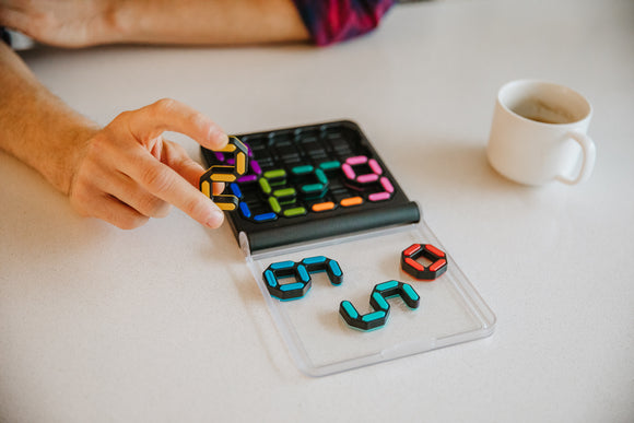 IQ Digits mechanical puzzle by Smart Toys and Games displayed on a white table. A person's hand holds a vibrant black and yellow digit piece, preparing to place it on a grid with colorful numbers. Additional pieces are laid out in a clear container nearby, showcasing various shapes and colors. A white cup sits in the background.