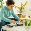 A young boy engages with the IQ Perplex mechanical puzzle from Smart Toys & Games. He is sitting at a table, concentrating as he arranges colorful, geometric puzzle pieces in a tray. An instruction booklet is open beside him, providing guidance for solving the puzzle. The setting is bright and inviting, with plants in the background, enhancing the play experience.