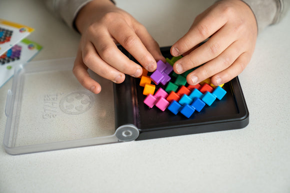 A close-up of a child's hands arranging colorful plastic pieces on a black base, part of the IQ Perplex mechanical puzzle by Smart Toys & Games. The pieces are in vibrant colors including purple, red, orange, and blue, showcasing an engaging and challenging design that encourages spatial reasoning and problem-solving skills.