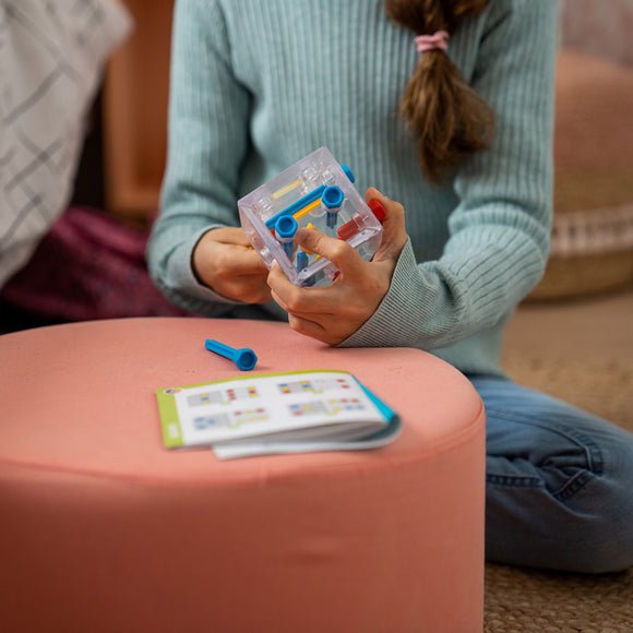 A child holding the Criss Cross Cube mechanical puzzle by Smart Toys & Games, with blue connectors and a guide booklet on a pink ottoman. The child is focused on solving the puzzle, surrounded by a cozy indoor setting.
