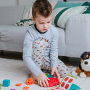 A young boy sitting on a rug, focused on the Apple Twist mechanical puzzle from Smart Toys & Games. He is wearing fox-patterned pajamas and using a red puzzle piece, surrounded by colorful pieces and instruction cards. A plush dog toy is nearby.