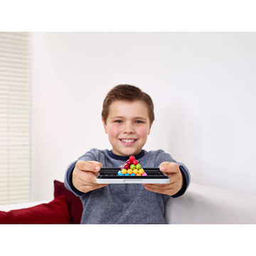 A young boy smiles while holding the IQ Puzzler Pro by SmartGames, showcasing colorful puzzle pieces arranged in a triangular formation on a black base. The background features a light-filled room with a bright window and a red sofa cushion.
