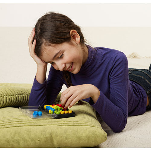 A young girl in a purple long-sleeve shirt is lying on a green cushion while concentrating on solving the IQ Twist mechanical puzzle by SmartGames. The puzzle features colorful pieces and a clear base, showcasing her engagement and joy in problem-solving.