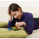 A young girl in a purple long-sleeve shirt is lying on a green cushion while concentrating on solving the IQ Twist mechanical puzzle by SmartGames. The puzzle features colorful pieces and a clear base, showcasing her engagement and joy in problem-solving.