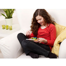 A young girl sitting comfortably on a couch, deeply engaged in playing with the IQ Twist - Multi-Level Logic puzzle by SmartGames. She has brown curly hair and is wearing a red long-sleeve shirt. The puzzle is an intricate mechanical challenge featuring colorful pieces that the girl is manipulating with both hands. The living room setting includes a plant in the background and a cozy atmosphere.