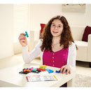 A young girl with curly hair sitting at a table, holding a colorful puzzle piece from the SmartGames Quadrillion mechanical puzzle. She is smiling and surrounded by various puzzle pieces in bright colors, with a solution booklet in front of her on the table.
