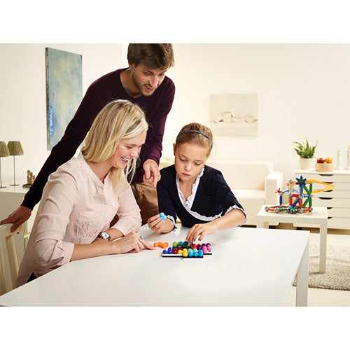 A family playing Quadrillion, a mechanical puzzle by SmartGames. The mother and daughter are engaged in placing colorful pieces on the table, while the father observes and provides guidance. The bright, inviting room features modern decor, emphasizing a family-friendly atmosphere for interactive play.