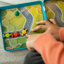 A child is playing with the Pole Position mechanical puzzle by Smart Toys & Games. The puzzle features a colorful road layout with three toy cars in vibrant colors (green, red, yellow) being maneuvered around the track. The child, wearing an orange sweater, is focused on directing the vehicles while seated on a soft surface.