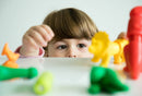 A young child curiously reaching out to play with colorful dinosaur-shaped toys. The child is focused on the vibrant green, yellow, orange, and red figures on a smooth surface, emphasizing a sensory play experience with engaging textures and forms.