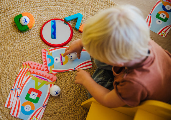 A young child interacts with the Smartmax My First Acrobats toy, featuring colorful animal figures and vibrant wooden pieces arranged on a textured mat. The child places a white ball with a smiley face onto a card depicting acrobat designs, aimed at developing fine motor skills and creativity through playful exploration.