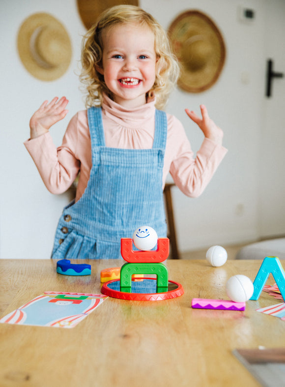 A joyful young girl enjoys playing with Smartmax My First Acrobats. Colorful stacking pieces and a smiling acrobat ball are on a wooden table, with an engaging atmosphere in the background.