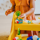 A young child is playing with the Smartmax Roboflex Large on a yellow table. The child is assembling colorful, flexible parts, including a blue box with eyes, a green stick, and various connecting tubes. The setup encourages creativity and imaginative play.