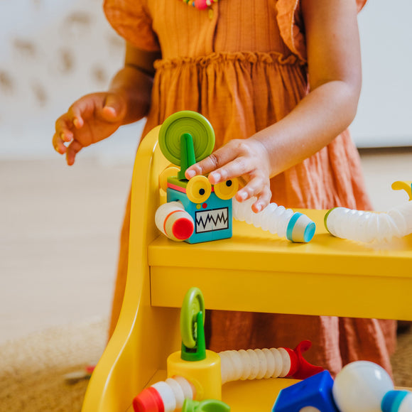 A young child is playing with the Smartmax Roboflex Large on a yellow table. The child is assembling colorful, flexible parts, including a blue box with eyes, a green stick, and various connecting tubes. The setup encourages creativity and imaginative play.